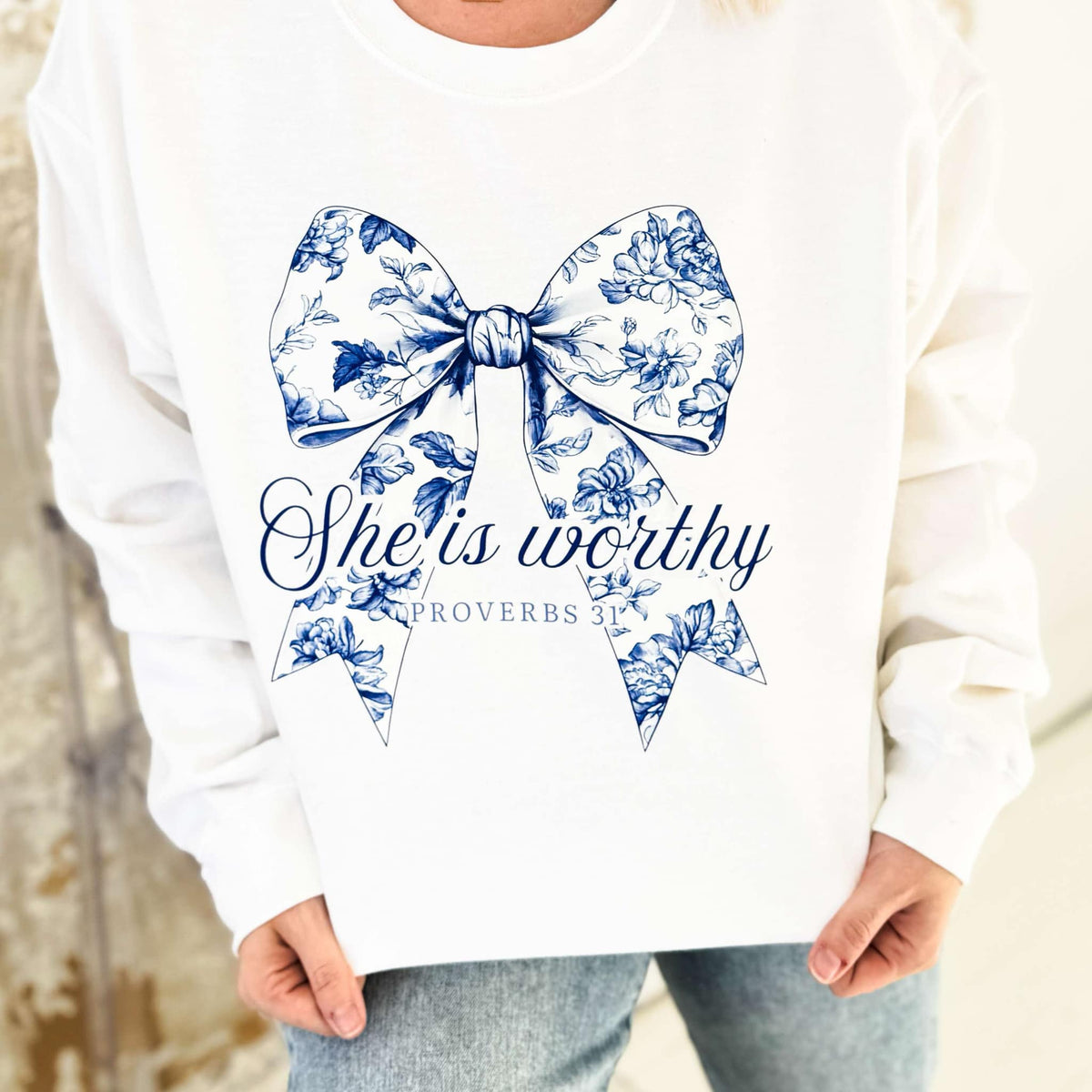 Women’s blue bow graphic sweatshirt by Envy Stylz Wholesale featuring "She Is Worthy" design on a white Gildan sweatshirt.