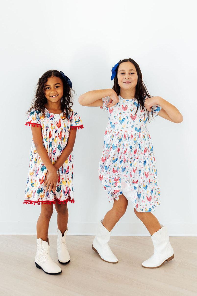 Mila & Rose rainbow rooster print short-sleeve twirl pocket dress for babies, featuring colorful rooster pattern and comfortable fit.